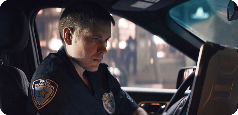 An officer at the wheel of a patrol car at night, lit by the screens around him.